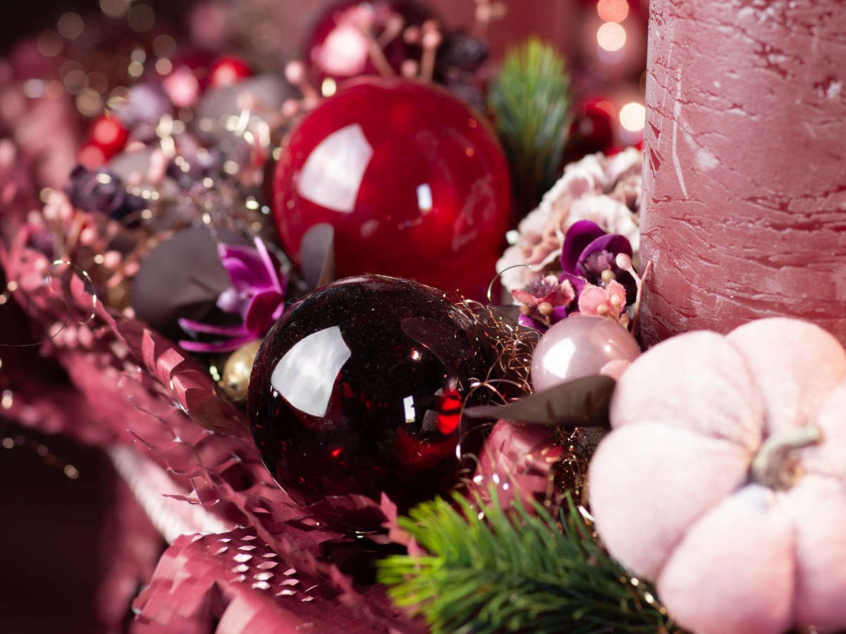 Adventsgesteck mit glänzenden Weihnachtskugeln in Rot und Bordeaux, rosa Kerzen und Samtkürbis